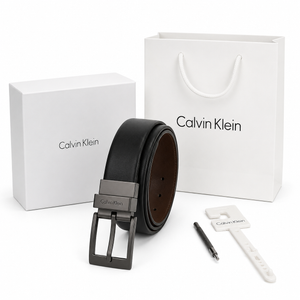 Complete product gift set display of the Calvin Klein mirror original reversible black leather belt rolled vertically next to its luxury box, branded shopping bag, and punch tool