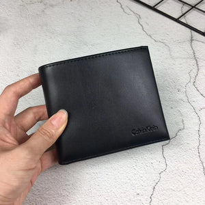 High-angle view of a closed Calvin Klein men's black leather bifold wallet resting on a white surface, highlighting the distinctive matte vertical center stripe and debossed logo
