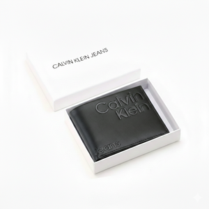 A closed Calvin Klein Jeans men's black genuine leather wallet featuring a large debossed logo, placed elegantly next to its matching closed white branded gift box