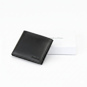 Top-down angled view of a Calvin Klein men's black smooth leather wallet with a small debossed logo, placed next to its matching closed white gift box