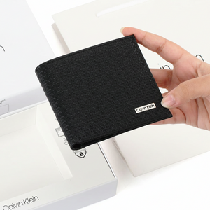 Hand holding a Calvin Klein men's black leather wallet featuring an embossed CK pattern and metal logo, displayed above its open white original gift box