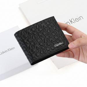 Hand holding a Calvin Klein men's black leather wallet showcasing the textured CK monogram pattern and metal plaque, presented in front of its original white branded packaging