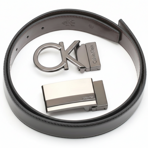 Angled perspective of a rolled Calvin Klein reversible leather belt and its two premium metal buckles, showcasing the high-quality craftsmanship and designer hardware