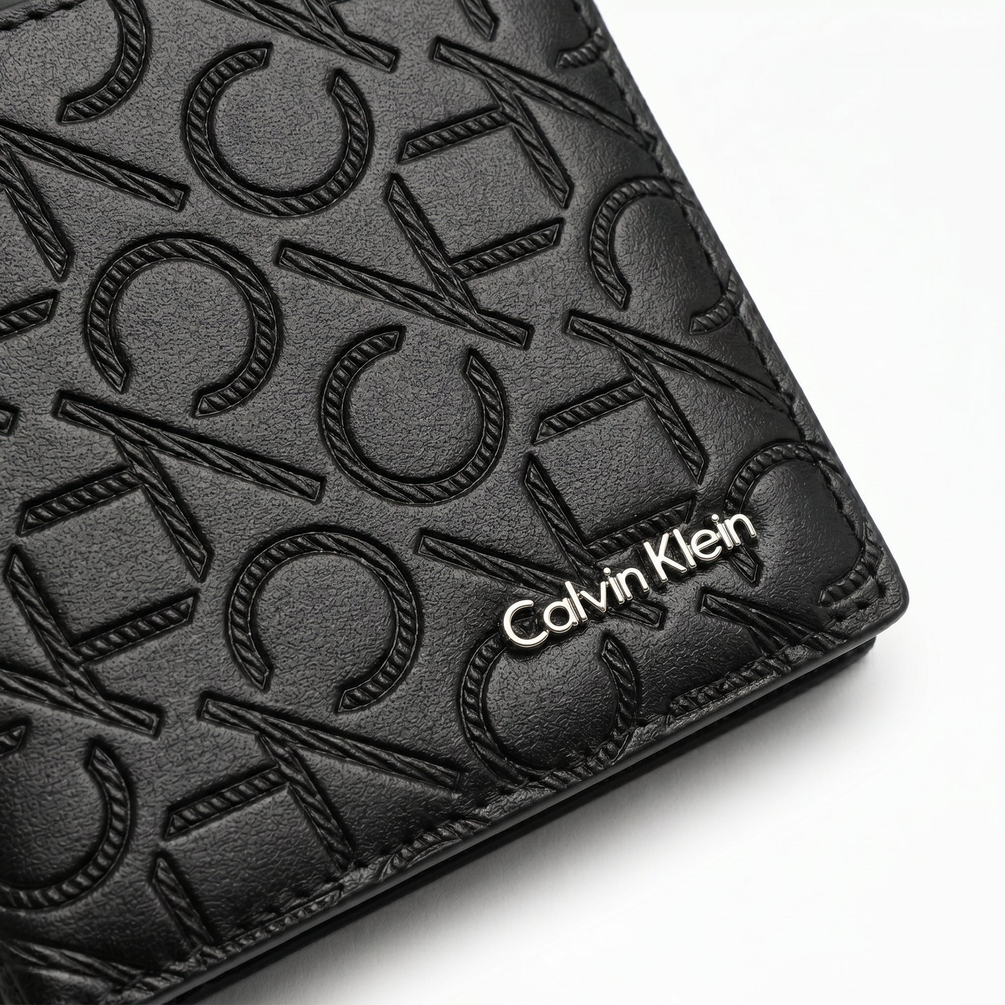 Macro close-up studio shot focusing on the engraved silver metal Calvin Klein logo plate and horizontal monogram fabric strip over black genuine leather