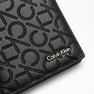 Macro close-up studio shot focusing on the engraved silver metal Calvin Klein logo plate and horizontal monogram fabric strip over black genuine leather
