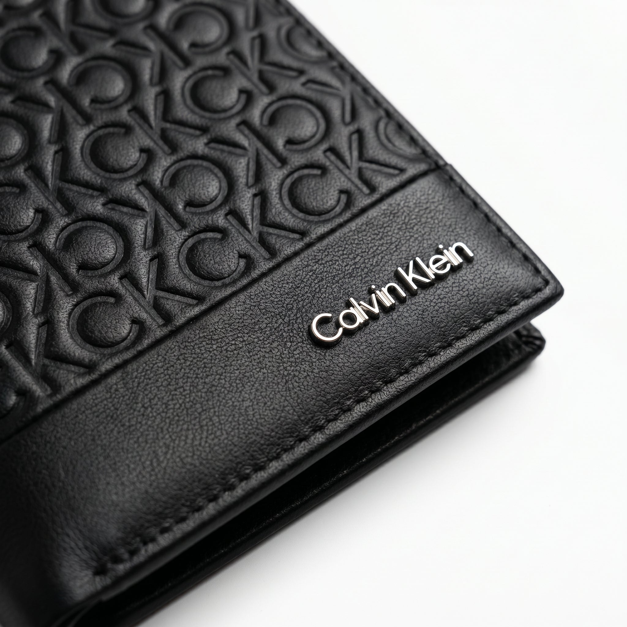 Macro close-up studio shot focusing on the polished silver Calvin Klein text logo over black genuine leather paneling integrated with embossed CK monogram pattern