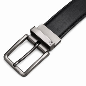 Live close-up smartphone photograph of the Calvin Klein reversible leather belt highlighting the silver rotatable buckle engraved with brand text over the black smooth side