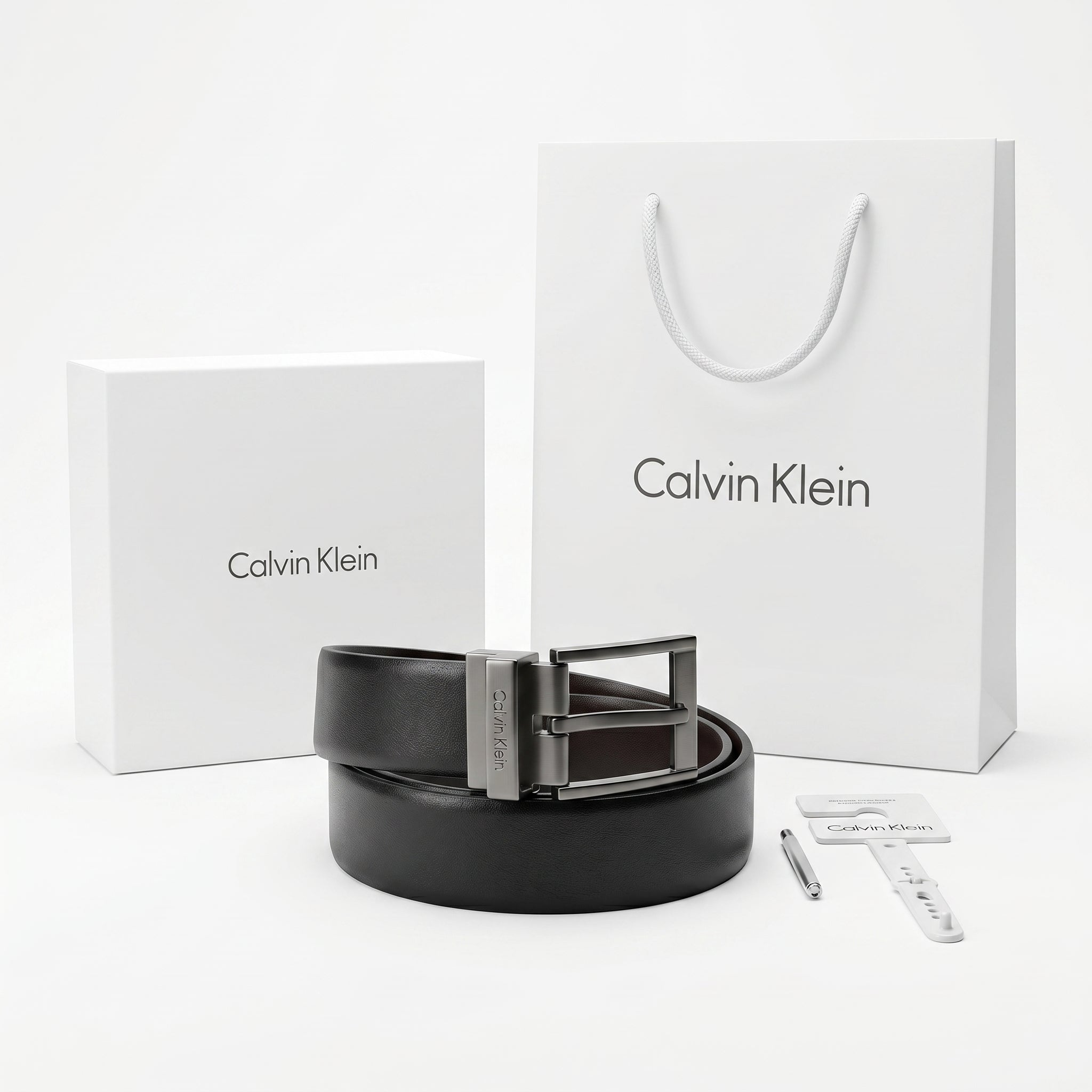 Full premium packaging presentation of the Calvin Klein mirror original reversible smooth black leather belt alongside its white gift box, matching shopping bag, tags, and hole punch tool