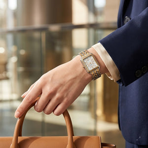 Hand holding a brown leather handbag with a person wearing a watch and part of a blue suit visible.
