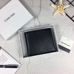 Hand holding a Calvin Klein men's black full grain leather wallet featuring a matte vertical stripe and small debossed logo, displayed above an open white gift box and shopping bag