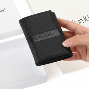 Hand holding a Calvin Klein men's black saffiano leather flap wallet over its original open white branded gift box, showing scale and premium packaging