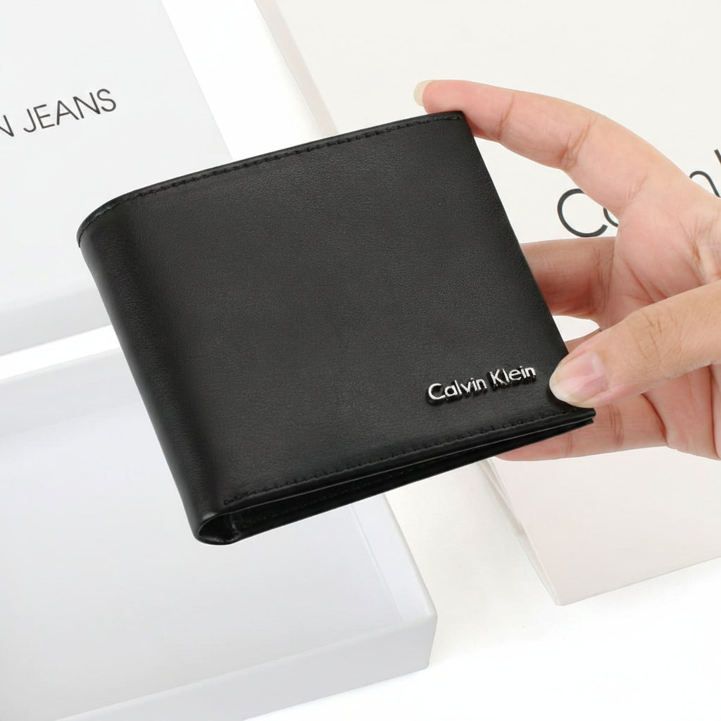 Hand holding a closed Calvin Klein men's black smooth leather wallet with a silver metal logo, displayed above its original open white branded gift box