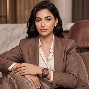 A woman in a brown blazer and cream shirt sits on a velvet chair, wearing a dark brown wooden watch with a round dial.