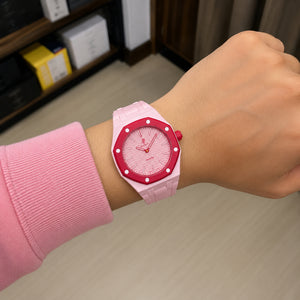 A close-up of a person's wrist wearing a pink and red watch with a light pink strap and a red bezel, set against an indoor background.