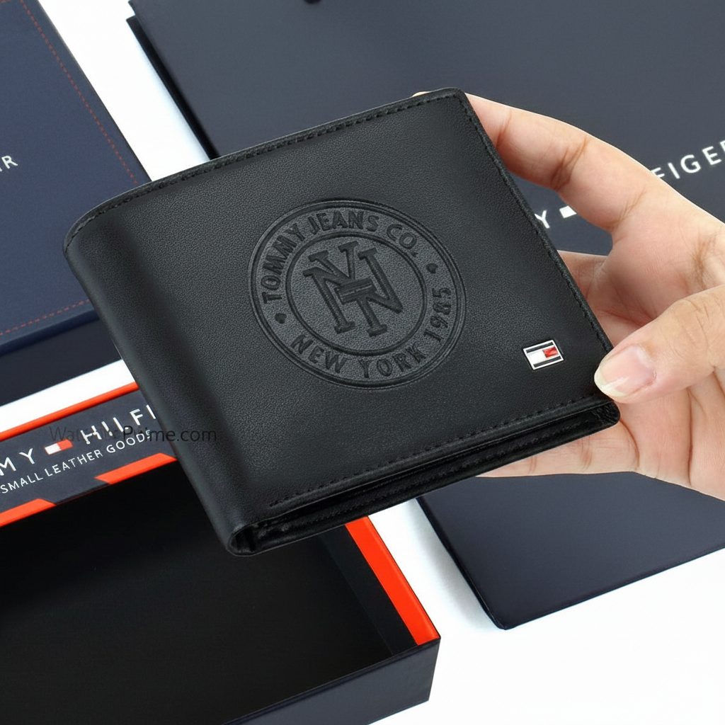 Hand holding Tommy Hilfiger black leather wallet showing real-life scale with embossed New York 1985 logo and gift packaging in background