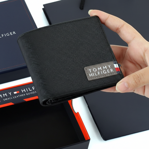 Handheld view of a Tommy Hilfiger black bifold wallet showcasing the durable Saffiano leather texture and the iconic red, white, and blue fabric branding