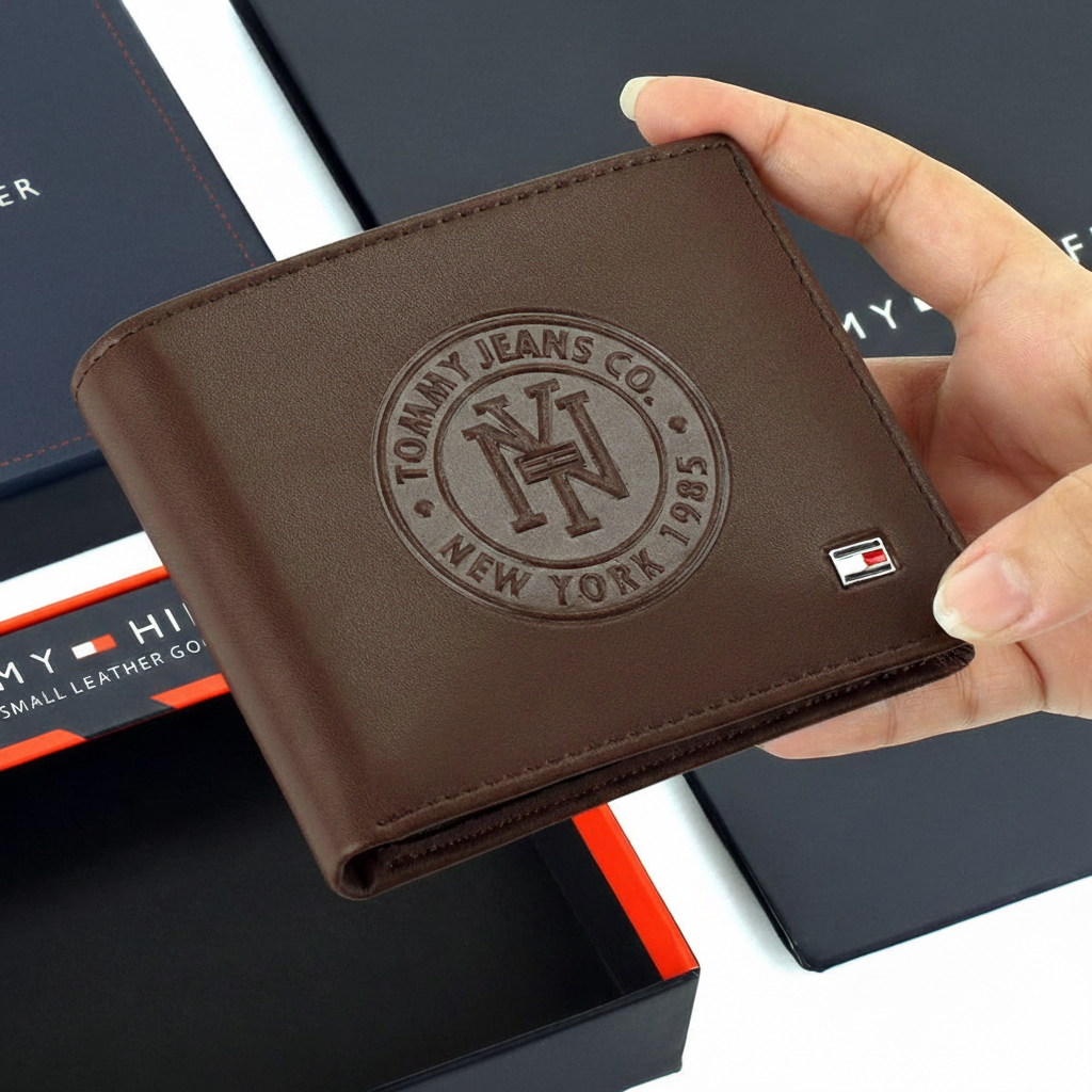 Hand holding Tommy Hilfiger brown leather wallet with large embossed heritage circle logo above navy blue gift box