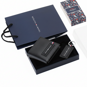 Elegant presentation of a Tommy Hilfiger men's black leather wallet and keychain set inside an open gift box, resting on a branded navy blue shopping bag with the Happy Holidays sleeve nearby