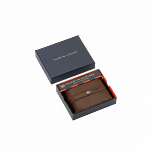 Handheld view of an authentic Tommy Hilfiger men's brown genuine leather bifold wallet with a sleek horizontal stripe and silver metal logo, shown with its premium navy gift box