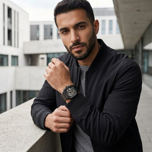 A man wearing a black smartwatch with a black strap and a black bomber jacket, leaning on a concrete balcony railing in an outdoor urban setting.