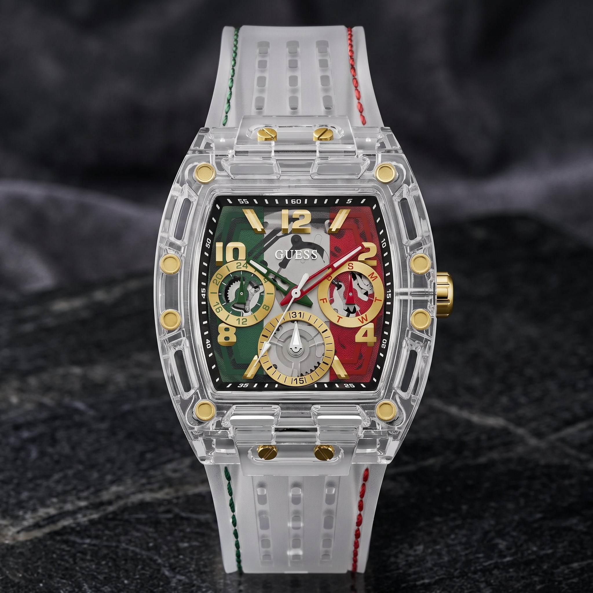 A luxury watch by Guess featuring a transparent acrylic case, a colorful dial with green, red, and gold sections, and a white silicone strap with green and red stitching, set against a dark textured background.