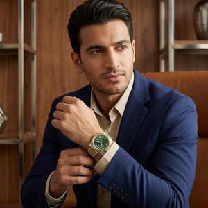 A man in a navy blue blazer and beige dress shirt wearing a gold-tone wristwatch with a green dial, posed in front of a warm wood-paneled background.