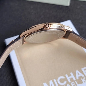 Close-up of the back of a Michael Kors watch with a rose gold case and light beige leather strap, resting on a branded cream-colored box.