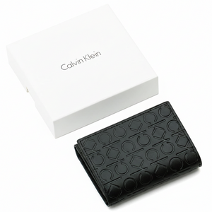 A closed Calvin Klein men's black leather flap wallet featuring an all-over embossed CK logo pattern, placed next to its matching closed white branded gift box