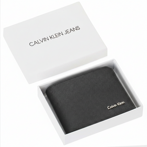 Top-down view of a Calvin Klein men's black saffiano leather wallet with a silver metal logo, presented inside its original white gift box next to a matching shopping bag