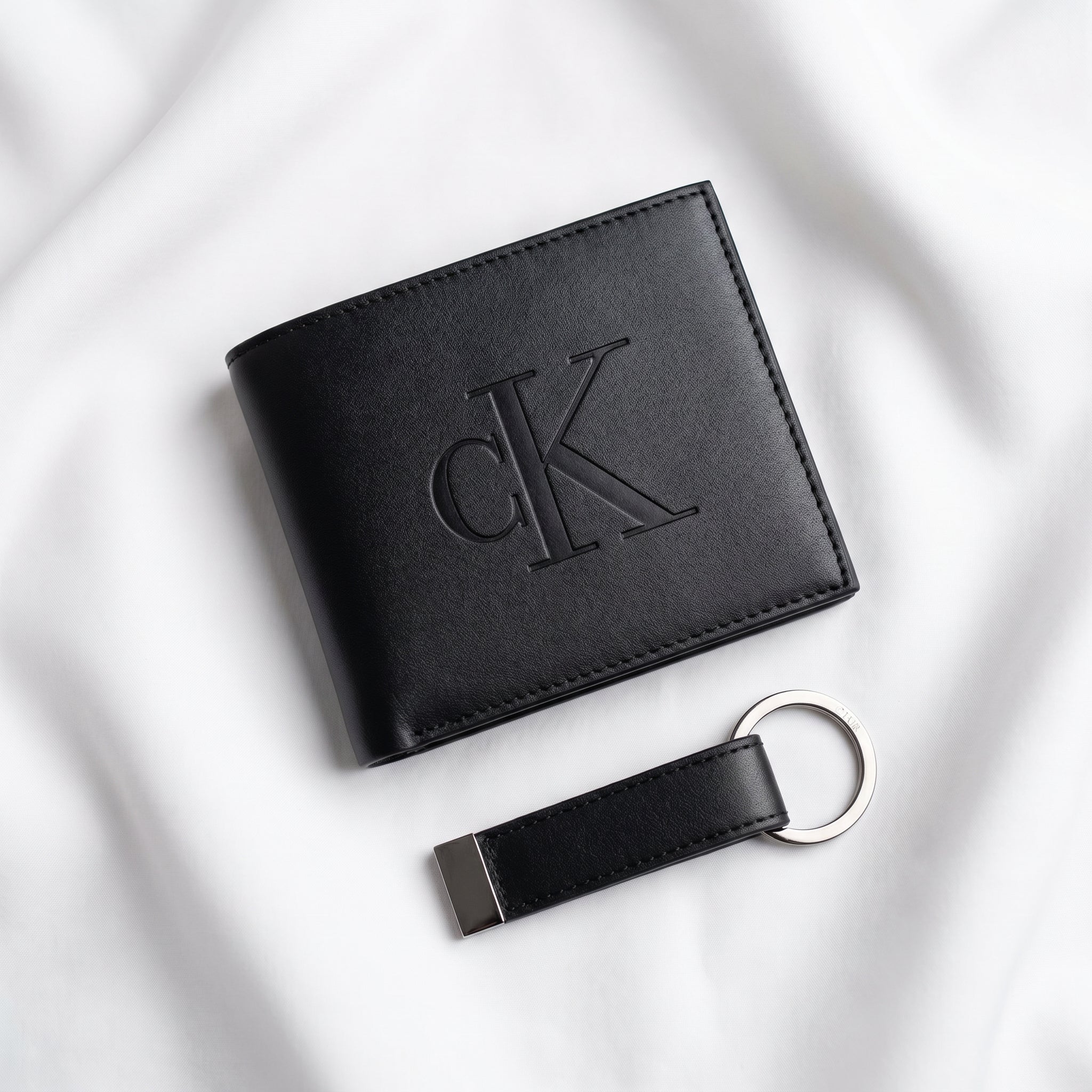 Studio product view of the Calvin Klein mirror original smooth black leather bifold wallet with large debossed center CK logo next to its matching standalone premium leather keychain