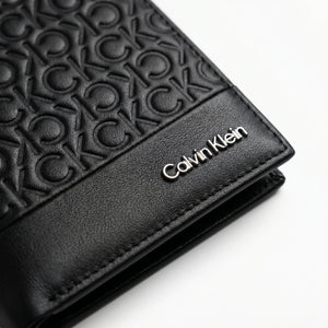 Macro close-up studio shot focusing on the polished silver Calvin Klein text logo over black genuine leather paneling integrated with embossed CK monogram pattern