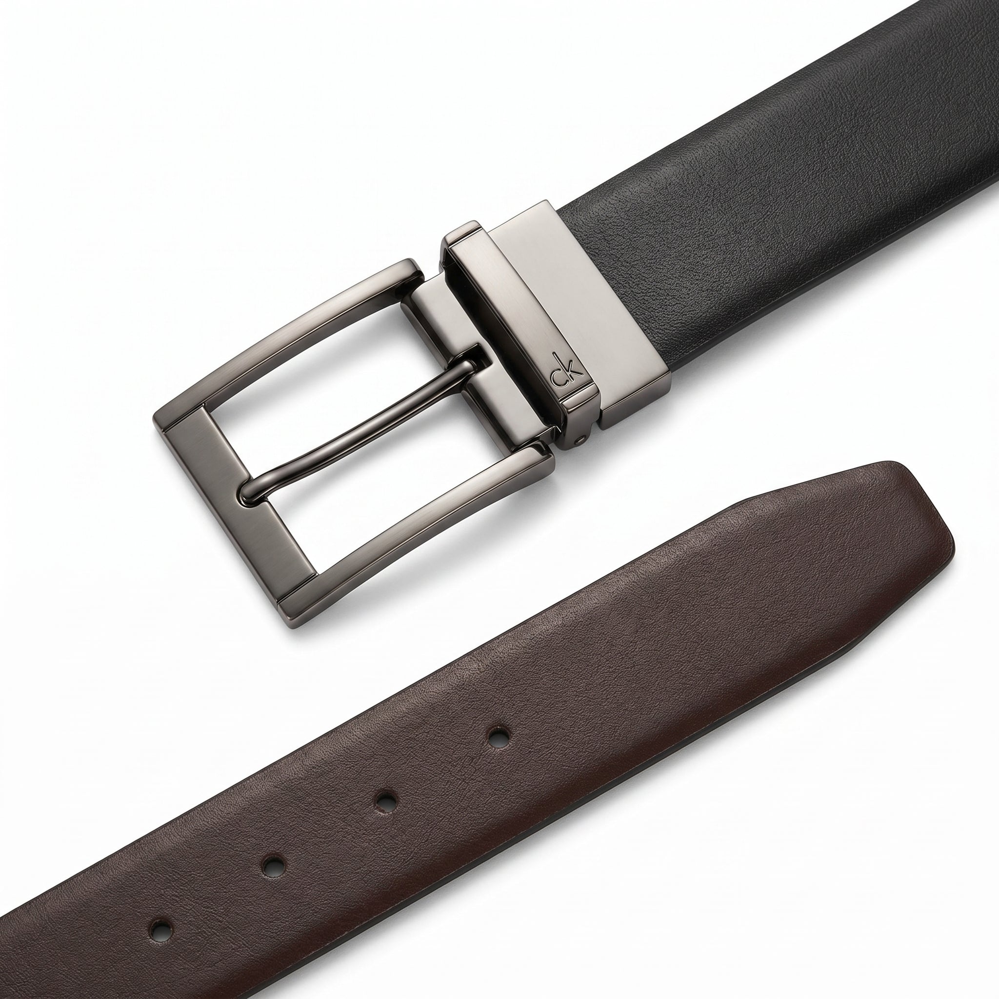 Macro close-up studio shot of the Calvin Klein mirror original reversible belt highlighting the rotatable gunmetal buckle with engraved ck logo alongside black and brown leather ends