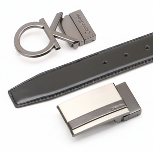 Detailed view of a Calvin Klein black leather belt shown with its two interchangeable designer buckles: a large cK plaque and a sleek rectangular metal buckle