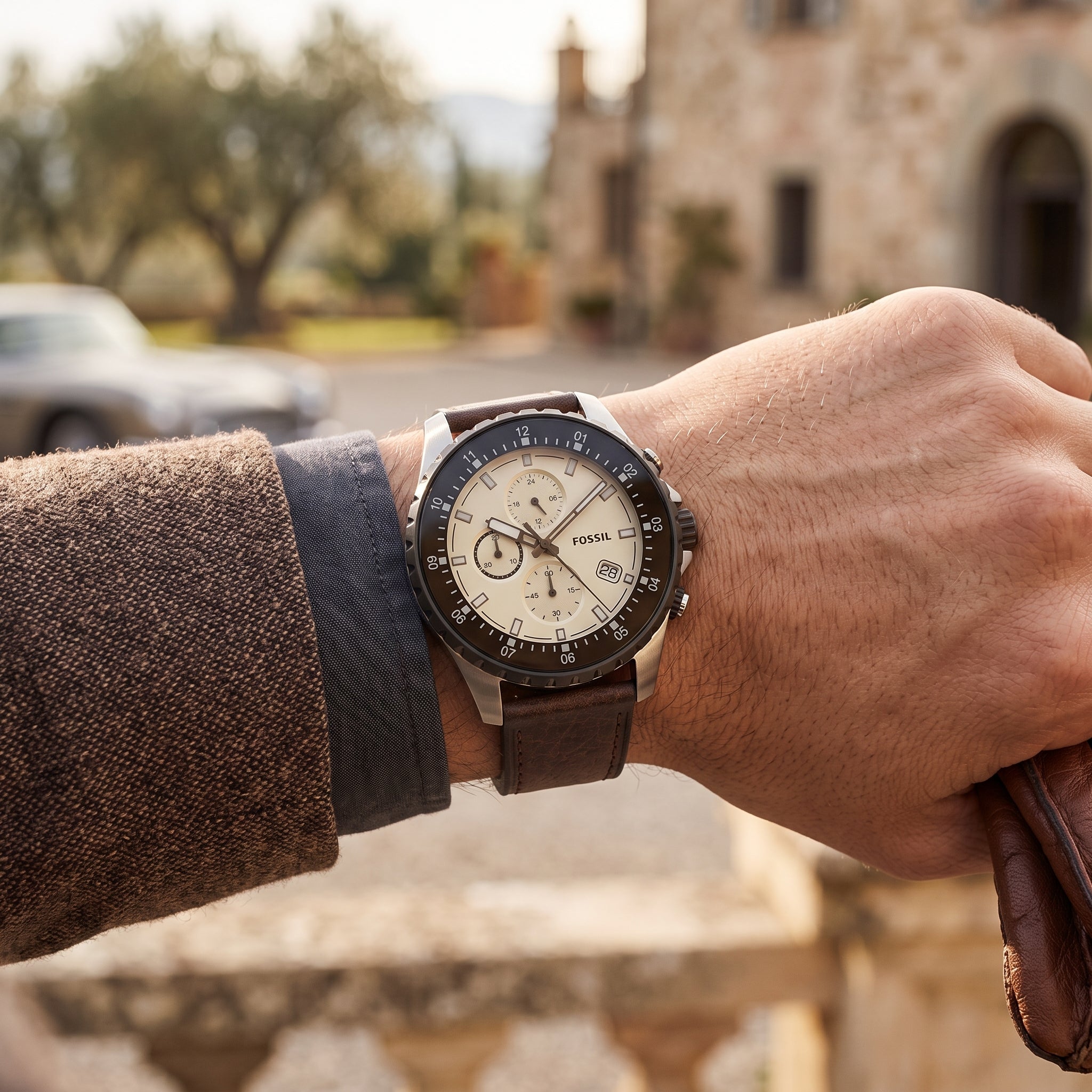 A close-up of a wrist wearing a Dial Corning chronograph watch with a cream dial, black bezel, and brown leather strap, set against a blurred outdoor background of a stone building and trees.