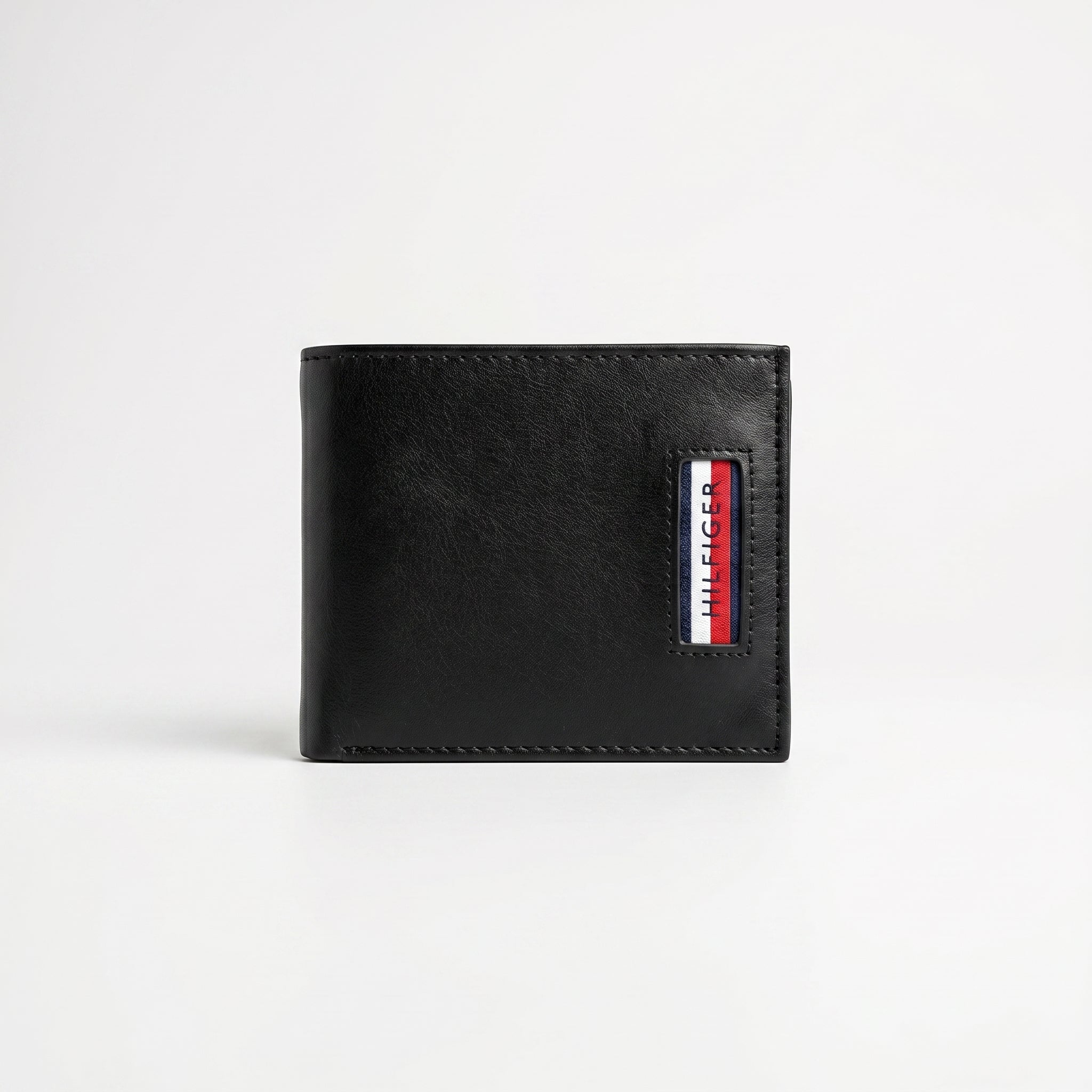 Straight front view product photography of the Tommy Hilfiger black trifold wallet showcasing the textured saffiano body, smooth leather corner, and metal flag logo