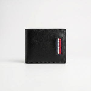 Straight front view product photography of the Tommy Hilfiger black trifold wallet showcasing the textured saffiano body, smooth leather corner, and metal flag logo