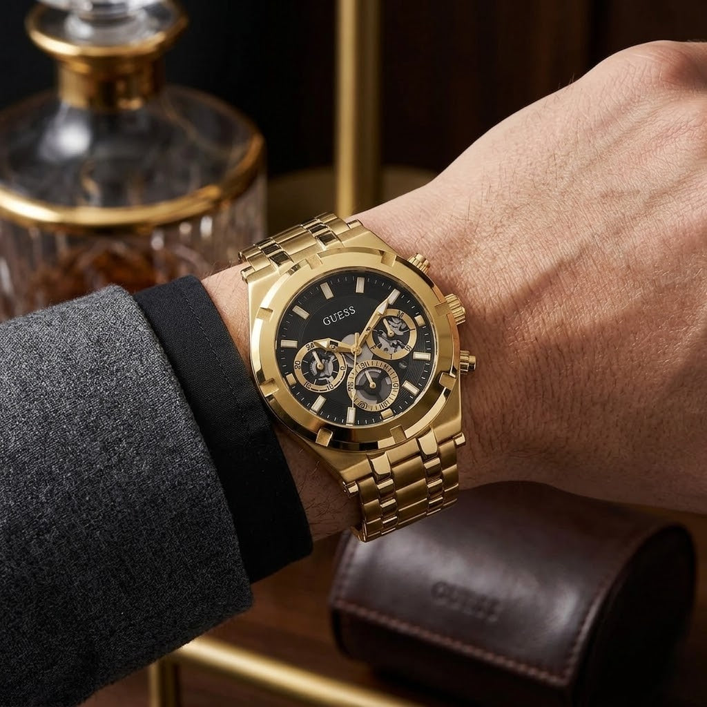 A gold-tone Guess wristwatch with a black dial and three subdials, shown worn on a person's wrist. The watch has a gold-tone metal link bracelet and case, with a black dial featuring gold-tone markers and hands.