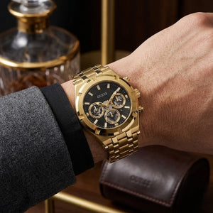 A gold-tone Guess wristwatch with a black dial and three subdials, shown worn on a person's wrist. The watch has a gold-tone metal link bracelet and case, with a black dial featuring gold-tone markers and hands.