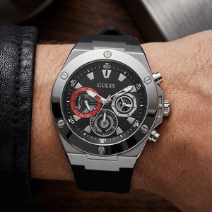 A Guess men's chronograph watch with a black dial, silver-tone case, and black rubber strap, shown worn on a wrist.