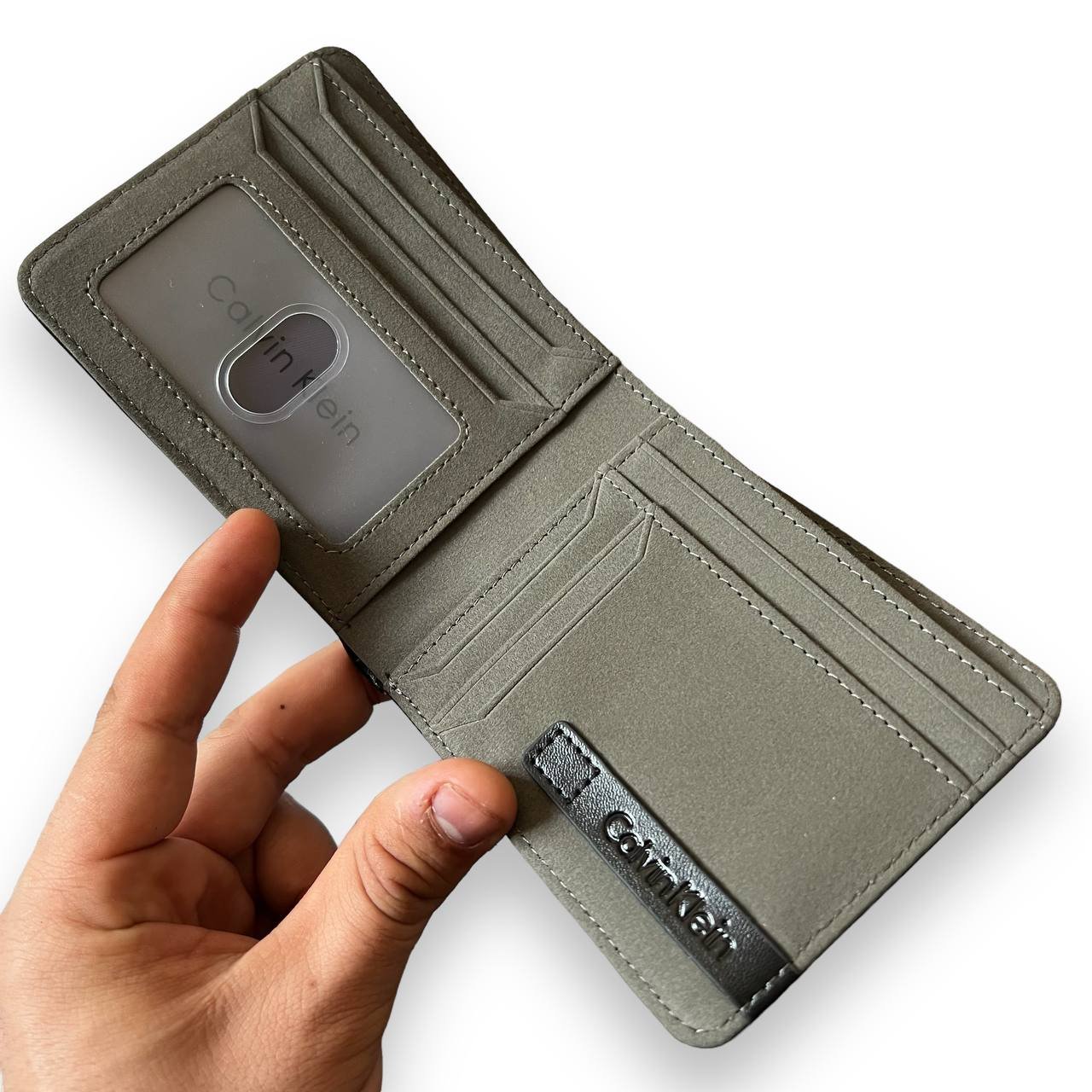 A hand holds open a Calvin Klein men's black leather bifold wallet, revealing the ID window slot.