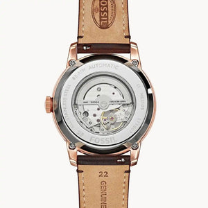 Fossil automatic watch with a transparent case showing the mechanical movement, finished in rose gold with a brown leather strap on a light gray background.