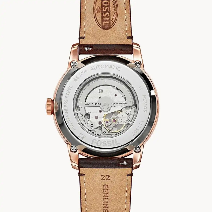 Fossil automatic watch with a transparent case showing the mechanical movement, finished in rose gold with a brown leather strap on a light gray background.