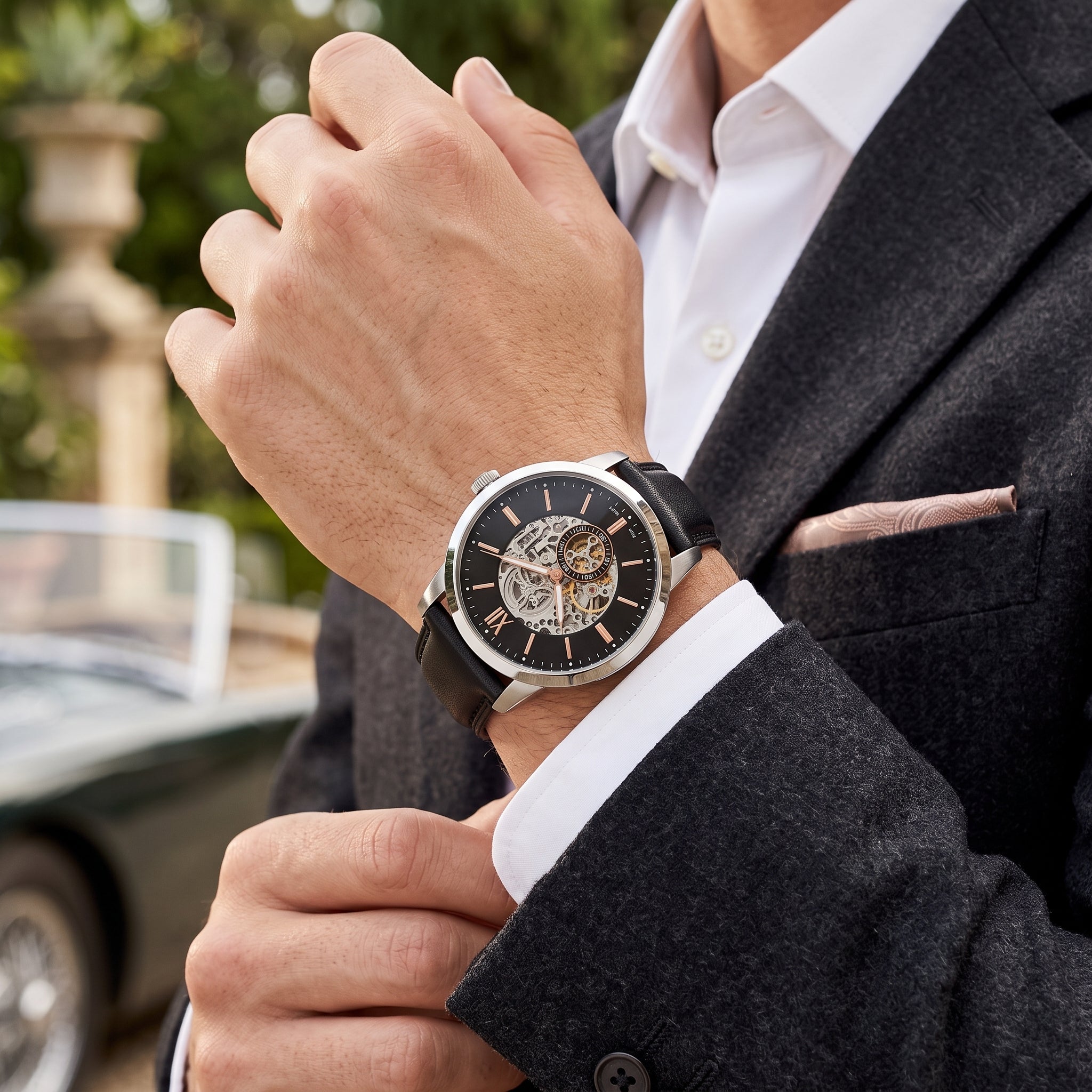 A close-up lifestyle shot of a man wearing a skeleton watch on his wrist, dressed in a dark gray suit jacket, white dress shirt, and pink pocket square, with a blurred car and garden background.