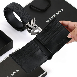 Hand holding open a Michael Kors men's black bifold wallet showcasing the interior card slots, with a reversible belt, MK buckle, and branded gift bag in the background