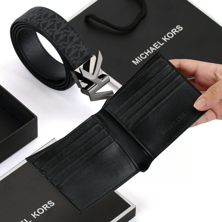 Hand holding open a Michael Kors men's black bifold wallet showcasing the interior card slots, with a reversible belt, MK buckle, and branded gift bag in the background