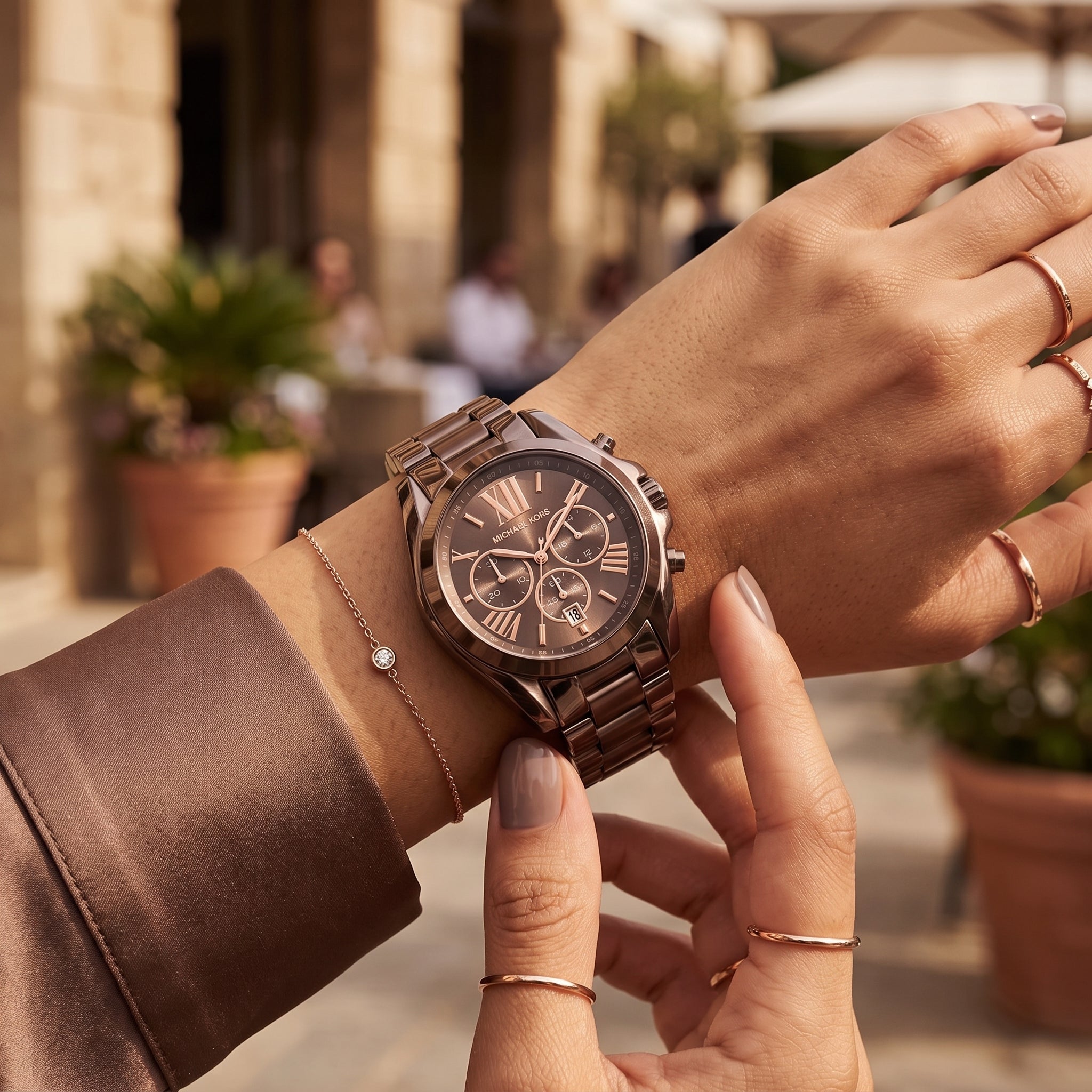 A close-up lifestyle shot of a woman's wrist wearing a rose gold chronograph watch with a matching metal link bracelet, set against a blurred outdoor cafe background.