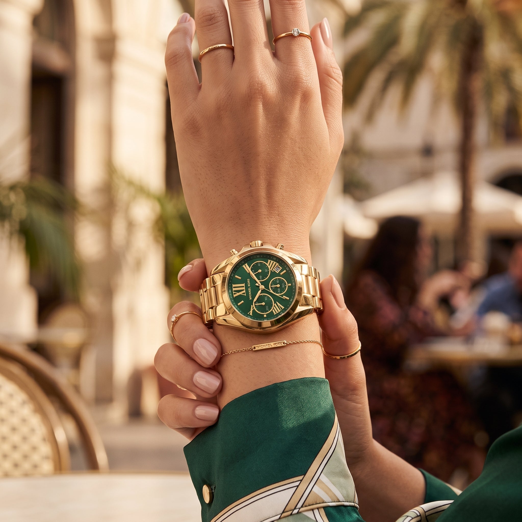A gold-tone chronograph watch with a green dial and matching green bracelet, shown worn on a person's wrist in an outdoor cafe setting.