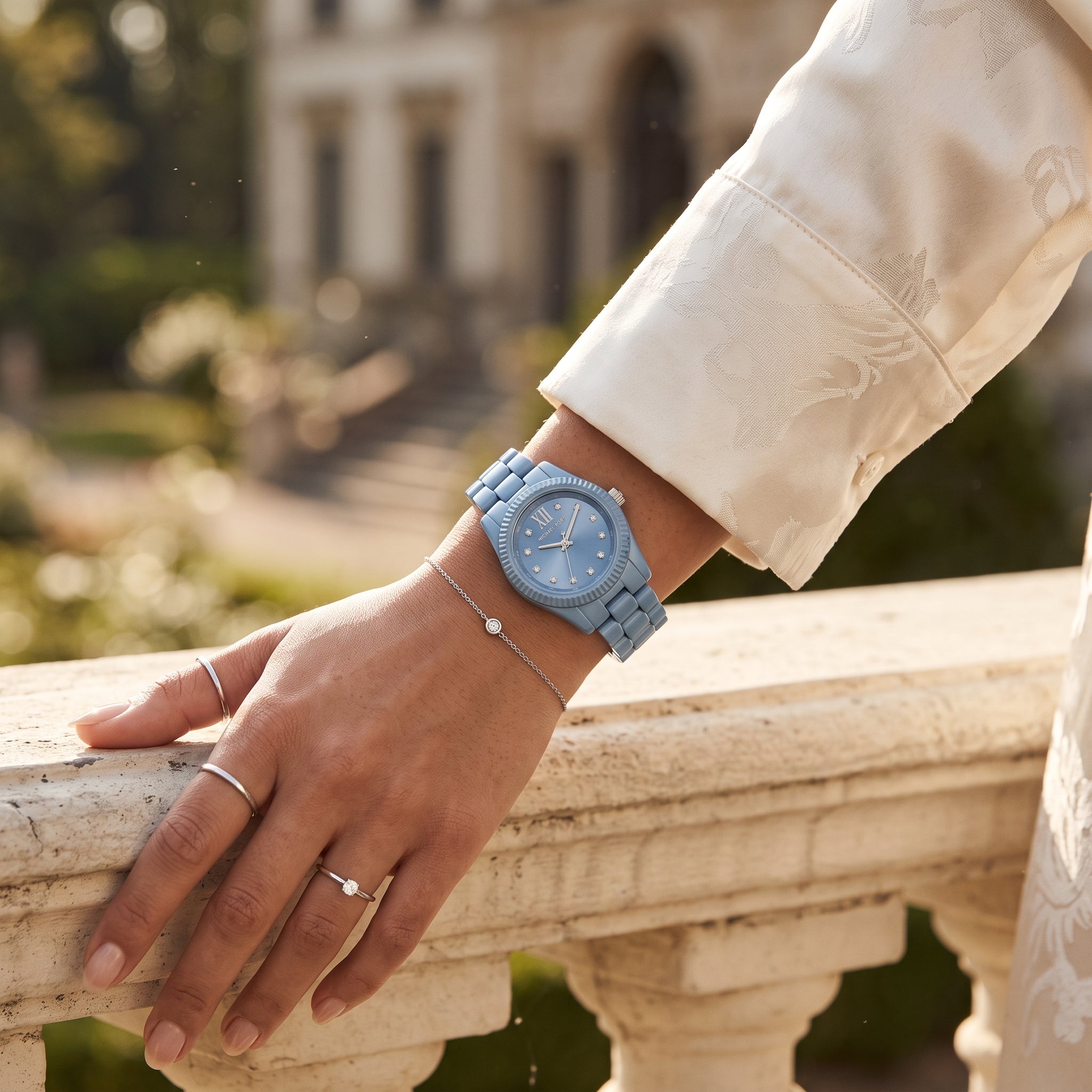 A person wearing a light blue wooden watch with a matching blue dial and bracelet, shown resting on a stone railing in an outdoor garden setting.