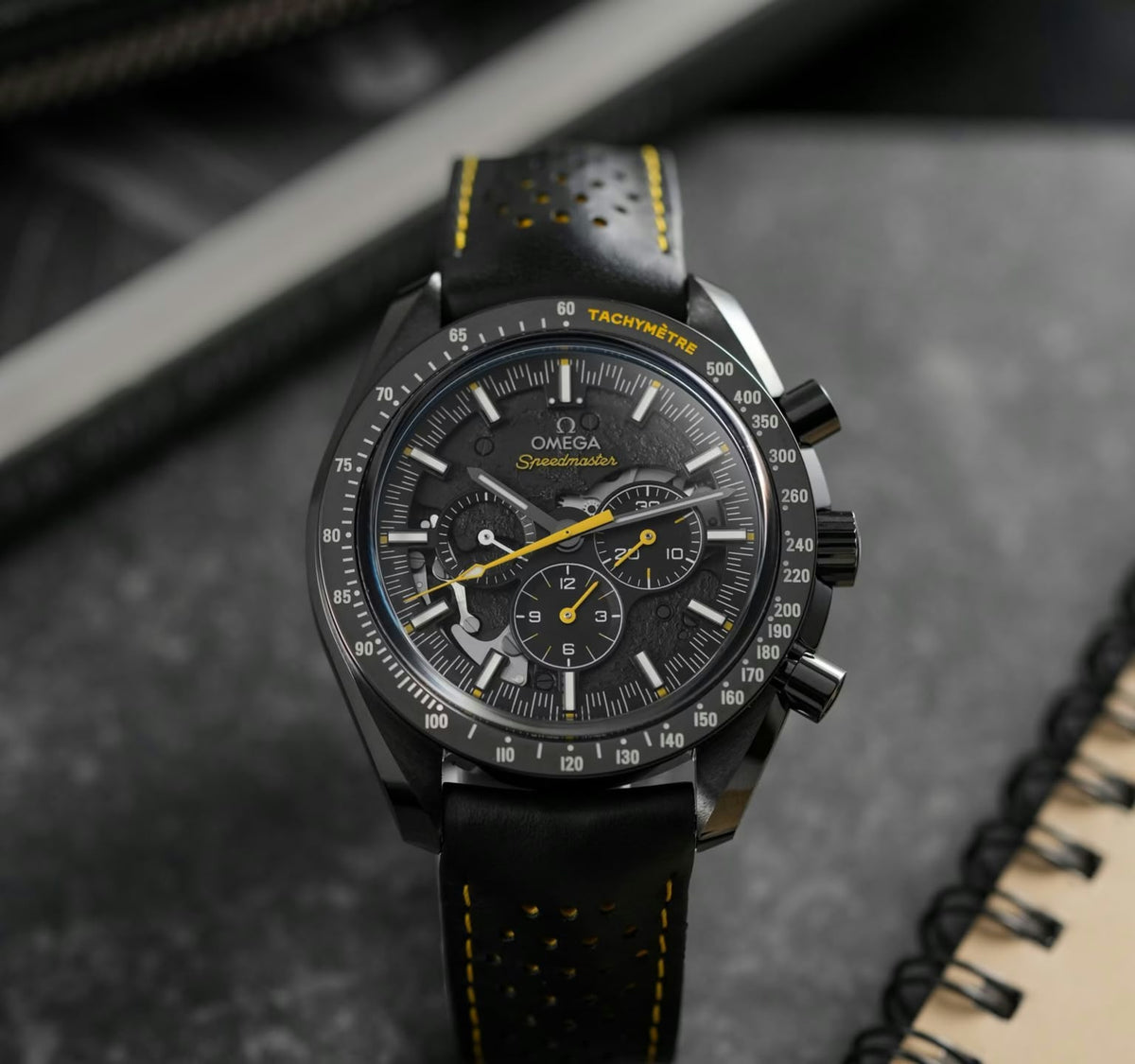 Omega Speedmaster Dark Side of the Moon Style men's chronograph watch, 45mm black case, black dial with yellow accents, black leather strap – angle view on dark background