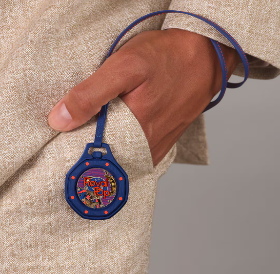 A close-up of a person's hand holding a small round blue pendant or tag with a colorful illustrated center and a blue strap, worn against a beige textured garment.